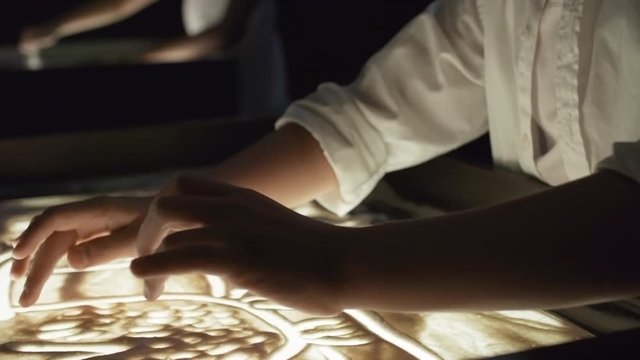 Tilt down shot of adorable little Asian girl using her fingers and drawing with sand on light table in dark classroom