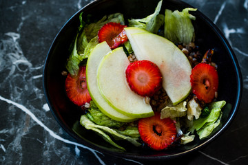 Apple Salad with Strawberry Slices, Buckwheat and Green Leaves in Black Bowl on Dark Granite Surface.