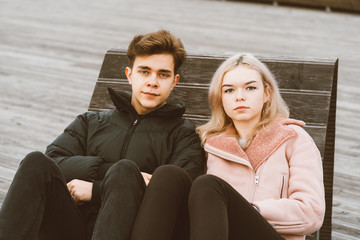 Loving teenagers are sitting on park bench in autumn, looking straight ahead. Teenage love concept. Cute brunette guy in dark jacket, beautiful young blonde girl in light pink jacket