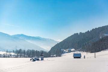 Winter landscape with a snow-covered field, trees and mountains in the background. Blue sky and a road leading to a small chapel. Schladming, Austria, Europe