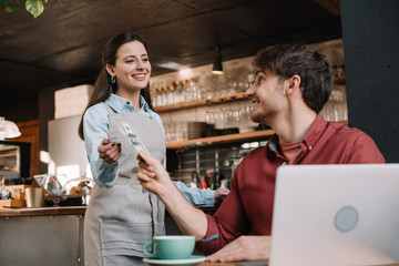 freelancer with laptop giving cash to smiling waitress in coffee shop