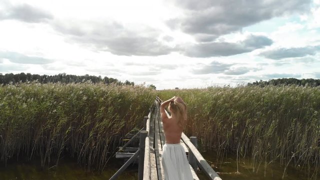 Aerial drone: Tender blonde woman with naked back walks in a field of wheat on whater wearing nothing but a white skirt
