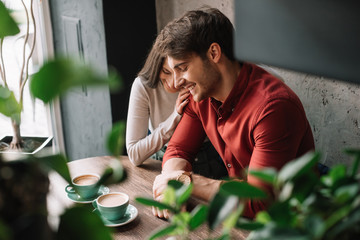 selective focus of green leaves and smiling romantic young couple hugging and drinking coffee in coffee shop