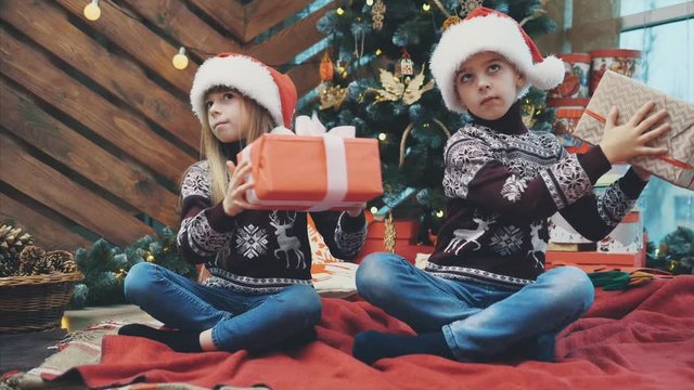 Little twins sitting under christmas tree, shaking present boxes, listening to and finding out what is inside.