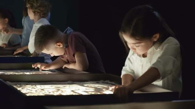 Tracking shot of serious little boy standing by light table and looking at camera during group sand animation class as young female art teacher talking to other children