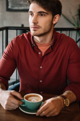 serous young man sitting at wooden table with cappuccino in coffee shop