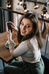 smiling young woman sitting at wooden table with cappuccino and using smartphone on balcony in coffee shop