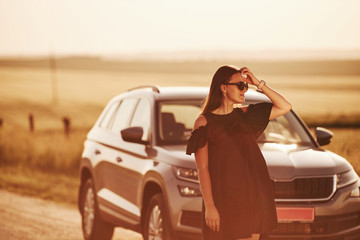 Looks far away. Girl in black clothes posing near the modern luxury automobile outdoors