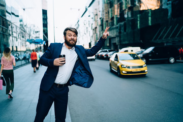 Handsome businessman with takeaway cup raising hand to call yellow cab on New york avenue,...