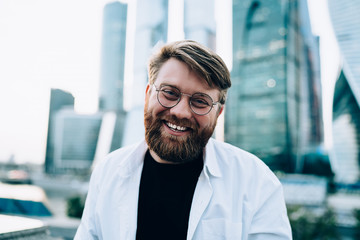 Close up portrait of cheerful Caucasian male in trendy optical spectacles for provide eyes protection smiling at camera during leisure time at city urban setting, happy man rejoicing at street