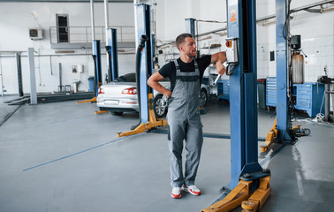 Looks at switch. Man in black shirt and grey uniform stands in garage after repairing broken car