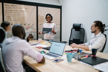 Cheerful afro american female financial expert holding paper with graphic report and explaining strategy for management project to employees sitting at meeting table, concept of brainstorming