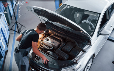 Under the hood. Man in grey uniform repairs white automobile indoors