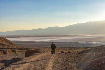 Lone hiker in shorts walking on a dusty gravel trail holding a water bottle in the desolated desert landscape of Death Valley, California, US. Mountains and a dried up lake at the background