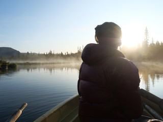 Fisherman man in rowing boat at lake Sjabatjakjaure in Beautiful sunny morning haze mist in Sweden Lapland nature. Mountains, birch trees, spruce forest. Blu sky and backlight