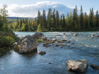 Beautiful northern landscape with azure blue river Kamajokk, mountains, boulders and spruce tree forest in Kvikkjokk in Swedish Lapland. Summer sunny day, white clouds
