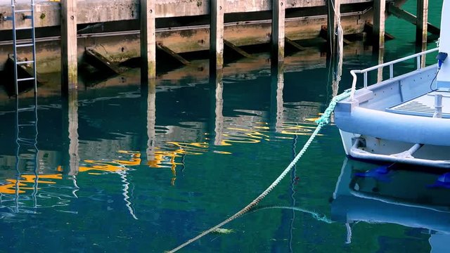 Reflection of wooden pier with ladder and small boat tied with rope are seen in calm waters in this serene clip.