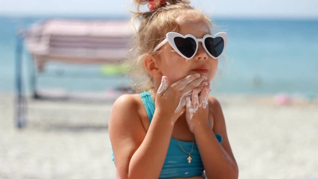 Child smearing sun cream fase. Sunburn. Suncream cream. Sunprotection cream. Cute baby girl in sunglasses sunbathes on a beach near the sea