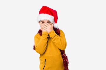 Little boy celebrating christmas day wearing a santa hat isolated blink through fingers frightened and nervous.