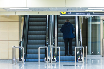 The alone man on the escalator or moving staircase with inscription departure in English and Chinese in the international airport or railway station from the back moving upstairs with luggage