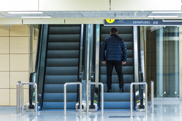 The alone man on the escalator or moving staircase with inscription departure in English and Chinese in the international airport or railway station from the back moving upstairs with luggage