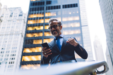 Joyful stylish businessman with fist up near handrail