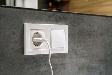 A closeup diagonal view of a group of white european electrical outlet with a plug inserted into it and a switch located on a concrete gray wall under a mirror in a modern bathroom