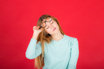 Portrait of a young red-haired beautiful woman of the 20s in a turquoise sweater, will take off glasses with closed eyes, isolated on a red background