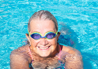 Close-up of a senior woman with swimming pool goggles in the transparent  water of the swimming pool. Smiles and happiness.. Healthy activity for pensioner.