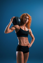 Stands with black soccer ball. Redhead female bodybuilder is in the studio on blue background