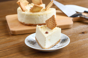 A piece of classic New York cheesecake decorated with square cookies on plate, on wooden table in coffee pastry shop with whole cake in the background. Cake sweet dessert for holiday, birthday party