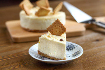 A piece of classic New York cheesecake decorated with square cookies on plate, on wooden table in coffee pastry shop with whole cake in the background. Cake sweet dessert for holiday, birthday party