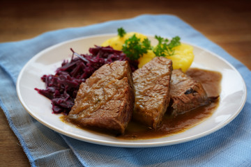 braised beef with sauce, potatoes and red cabbage, served on a white plate and blue napkin on a rustic wooden table