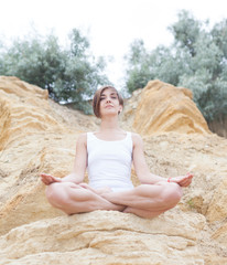 Fototapeta na wymiar A beautiful young girl with short hair is dressed in shorts and a white jersey is practicing yoga on the background of rocks. Pose of the lotus. The concept of calm and concentration