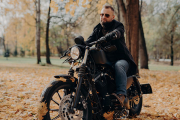 Obraz na płótnie Canvas Male motorcyclist drives in nature on fast bike, wears shades, black jacket, gloves, jeans and boots, enjoys autumn season, spends free time actively, ready for long trip. People, transport, driving