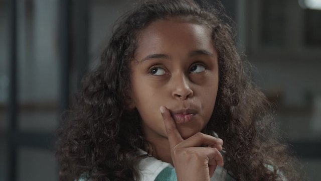 Cute pensive elementary age african american girl with puzzeled expression, pondering over something, looking upward in contemplation with forefinger on her lips, with placid and thoughtful look.