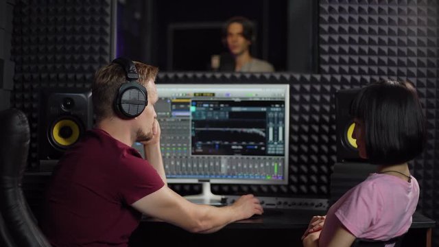 Back view zoom in of middle aged sound engineer and young female music producer looking at monitor in recording studio and discussing song being performed by singer in booth