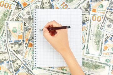 Hand with pen over blank note book on background of dollar bills and coins. Financial concept with copy space for your text. Flat layout, top view.