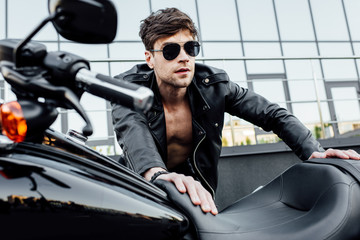 Fototapeta na wymiar selective focus of handsome man in sunglasses and black leather jacket leaning on motorcycle and looking forward