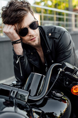 Fototapeta na wymiar handsome young motorcyclist in sunglasses holding head with hand and looking away while sitting on motorcycle