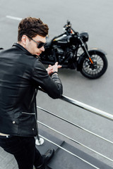 selective focus of man in leather jacket standing near metal fence with motorcycle on background