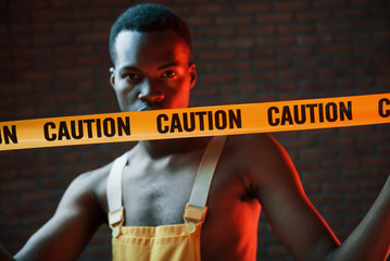 Yellow caution tape and uniform. Futuristic neon lighting. Young african american man in the studio