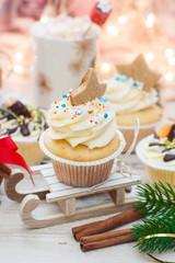 Christmas decorated cupcakes on wooden sledge. Cinnamon rolls, cotton flowers, tangerine bites, christmas tree branches and gingerbread cookies. String lights and cup with marshmallow on background.