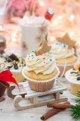 Christmas decorated cupcakes on wooden sledge. Cinnamon rolls, cotton flowers, tangerine bites, christmas tree branches and gingerbread cookies. String lights and cup with marshmallow on background.