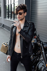 Fototapeta na wymiar young motorcyclist with muscular naked torso standing near black motorcycle smoking and holding lighter