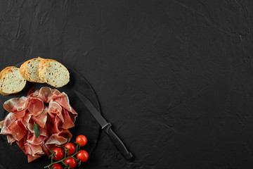 Sliced jamon, cherry tomatoes, baguette, herbs and knife on black stone slate board against dark grey background. Close-up. Top view. Copy space.