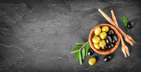Fresh olives in wooden bowl on dark stone table. Black and green olive with pickers or sticks from...