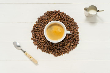 White cup with coffee in the pile of coffee beans on the white wooden background. Spoon, mug and  pitcher with milk. Food and drink background photography.