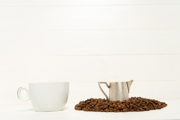 Pitcher with milk in the pile of coffee beans and cup of coffee on the white wooden background. Food and drink background photography.