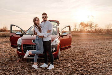 Naklejka premium Young Happy Couple Dressed Alike in White Shirt and Jeans Enjoying Road Trip at Their New Car, Beautiful Sunset on the Field, Vacation and Travel Concept